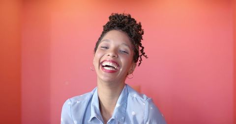 African american woman laughing and smiling in pastel pink studio wearing polka-dot blouse