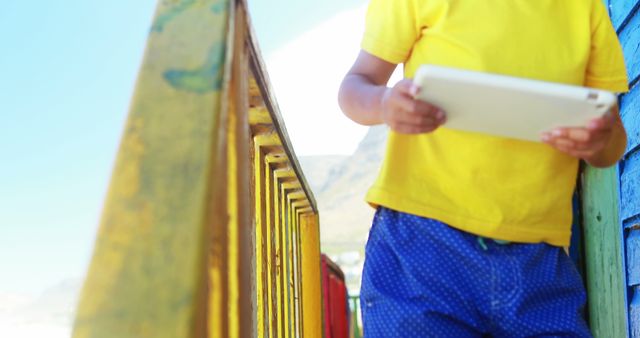 Child wearing yellow shirt and blue shorts holding tablet while standing near colorful wooden railing. Perfect for tech, summer, outdoor activities, and lifestyle contexts.