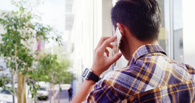 A young Asian man is engaged in a phone conversation outdoors, with copy space. His casual attire and smartwatch suggest a blend of technology with everyday life.