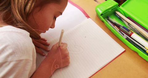 Chinese schoolgirl drawing in lined notebook at desk with green pencil case, markers