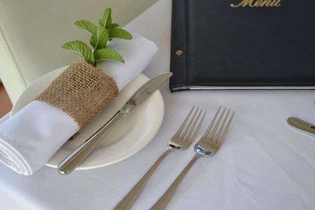 Elegant table setting featuring a neatly wrapped napkin in burlap with a sprig of mint. Includes silverware arranged properly on a white tablecloth next to a menu. Ideal for illustrating concepts of fine dining, restaurant preparation, and catering services.