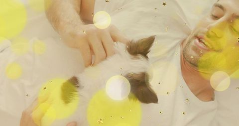 Smiling man cuddling black-and-white puppy on bed with dreamy yellow bokeh, cozy morning
