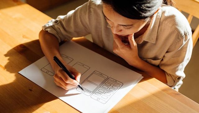 Woman sketching mobile app wireframes on paper at wooden table in warm sunlight for ux design