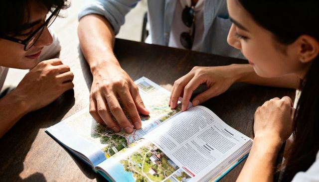 Young friends planning tropical trip tracing routes in guidebook at sunlit café