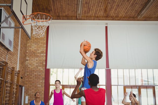 High school kids are actively playing basketball in a gymnasium. One player is jumping to shoot the ball towards the hoop while others are ready to rebound or defend. This image can be used for promoting youth sports programs, school athletic events, teamwork and fitness activities.