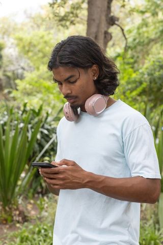 Young man standing in park wearing blue t-shirt and pink headphones, using smartphone