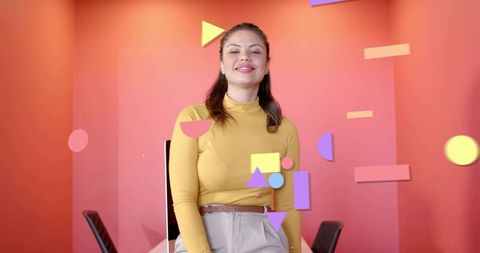 Smiling woman in coral studio wearing mustard sweater and beige pants with geometric shapes