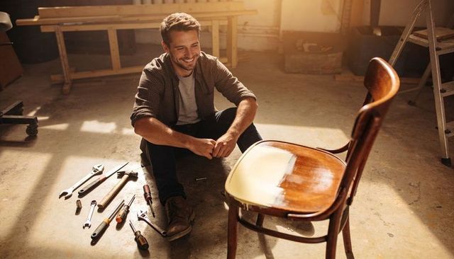 Smiling craftsman refinishing vintage wooden chair in sunlit workshop, tools on floor
