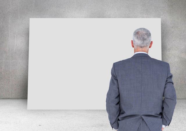 Middle-aged businessman in suit stands with gray concrete wall and large blank card. Useful for business presentations, marketing, corporate planning, decision-making processes, advertisements, and strategic planning imagery.