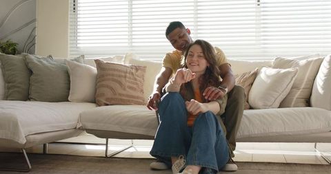 Diverse couple embracing on floor in cozy neutral living room with natural daylight
