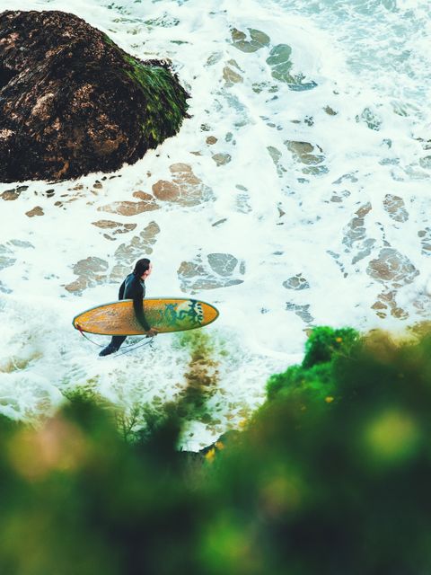The image shows a surfer holding a surfboard and walking into ocean waves from a rocky beach. This is perfect for content related to surfing, adventure sports, beach activities, and coastal lifestyles. Great for travel blogs, sports magazines, and holiday advertisements.