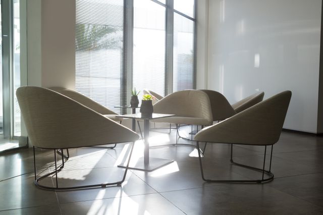 This image depicts a modern office lounge area with empty chairs arranged around a small table. Sunlight streams through large windows, creating a bright and welcoming atmosphere. Ideal for use in business presentations, office design inspiration, corporate brochures, and articles about modern workspaces.