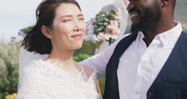 Image portrait of happy diverse bride and groom embracing and smiling to camera at outdoor wedding. Marriage, love, happiness and inclusivity concept.