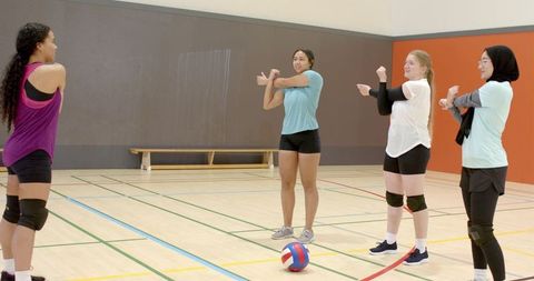 Diverse women's volleyball team warming up stretching arms on indoor court with ball