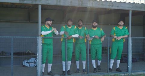 Five cricket players clapping and leaning on railing in sunlit dugout showing team unity