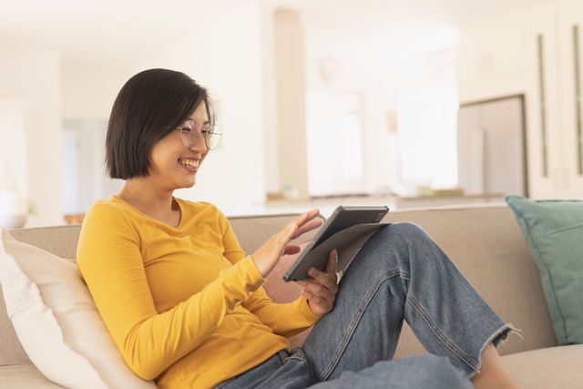 Asian woman sitting on a comfortable sofa in a bright living room, using a tablet and smiling. Ideal for content related to modern lifestyle, technology use at home, relaxation, and domestic life. Perfect for articles, blogs, or advertisements focusing on home comfort, digital devices, and leisure activities.