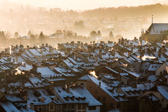 Snow-covered rooftops of a European town basked in the soft morning light during sunrise. Smoke rising from chimneys creates a cozy atmosphere. Hillside town with an expanse of traditional houses and a church visible through the morning fog. Ideal for use in travel blogs, winter-themed promotions, and tourism marketing materials.
