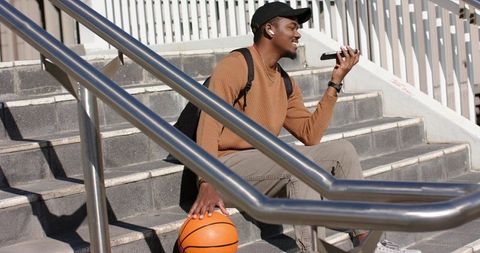 Young African American Student Sitting on Outdoor Steps Holding Basketball and Phone