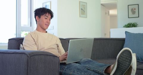 Asian man relaxing on sofa using laptop for remote work in bright modern living room
