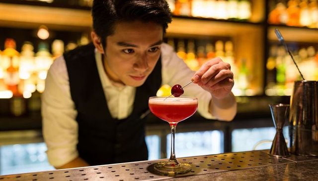 Bartender placing cherry garnish on frothy coupe cocktail in dimly lit bar
