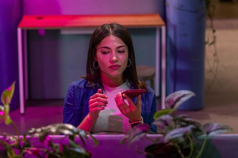 Young woman using smartphone in coworking lounge with magenta and blue ambient lighting