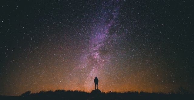 Silhouette of person standing on hilltop observing the star-filled night sky, with Milky Way galaxy glowing brightly. It captures the awe and wonder of the universe, perfect for backgrounds, astronomy events, wallpaper, or inspiring travel-related content, bringing feelings of serenity and fascination.