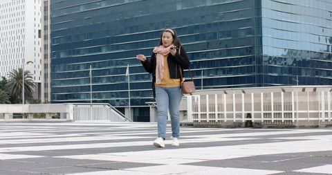 Mature woman walking and talking on smartphone across striped urban plaza with glass tower