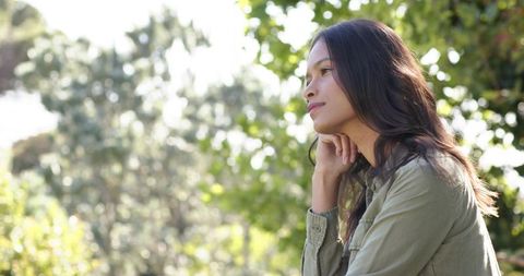 Asian woman gazing thoughtfully in sunlit park, resting chin on hand, serene nature moment