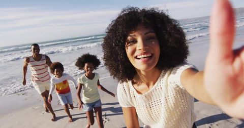 Smiling woman taking selfie with family running and laughing on sunny beach shoreline