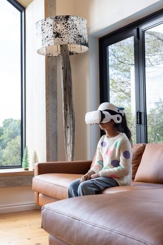 Asian child wearing vr headset sitting on tan leather sofa by window cozy modern tech play