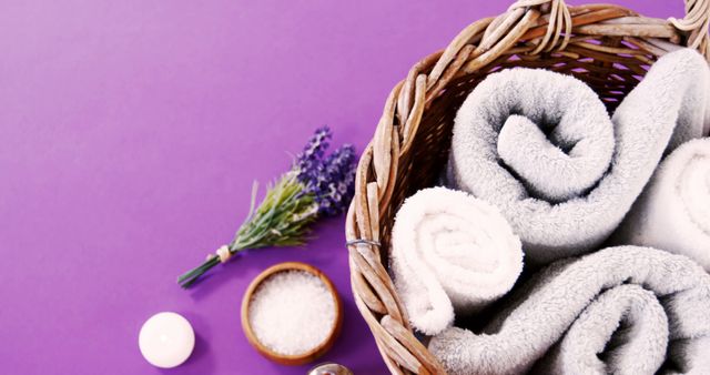Rolled gray towels are neatly arranged in a wicker basket next to a bundle of lavender and a bowl of bath salts on a purple background, with copy space. It conveys a sense of relaxation and spa-like tranquility, ideal for wellness and self-care themes.