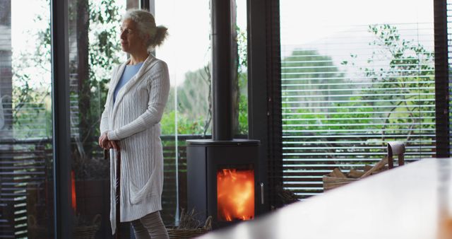 Elderly woman standing by a fireplace in a modern, cozy home. She appears thoughtful and peaceful, enjoying the warm ambiance. Useful for illustrating themes of retirement, comfort, and homely environments. Ideal for wellness, lifestyle blogs, or retirement planning advertisements.