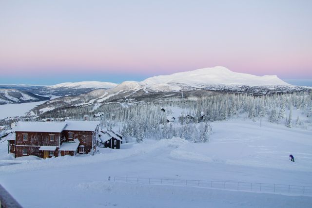 This image shows a peaceful snowy mountain village at sunrise during winter. The gentle pink hue of the sky contrasts with the white snow covering the landscape. Cabins and trees spread across the mountainside create a serene scene. This would be ideal for promoting winter tourism, holiday retreats, or showcasing scenic natural beauty.