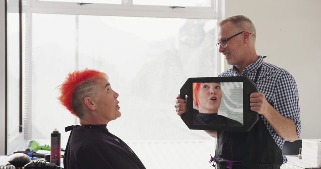 Hairdresser presenting finished haircut to middle-aged client with vibrant orange hair using a mirror in a modern barber shop. Client appears to be happily surprised. This can be used for content related to beauty, hair care, customer satisfaction, and personal grooming services.