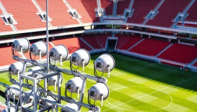 Floodlights overlooking striped soccer pitch and empty red stadium stands casting shadows
