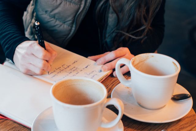 Woman is writing in a notebook while sitting at a coffee shop table with two coffee cups. Great for illustrating themes of productivity, studying, writing, and casual meetings in a cozy, relaxed environment. Can be used for blogs, articles, and advertisements related to coffee shops, writing, or freelance work.