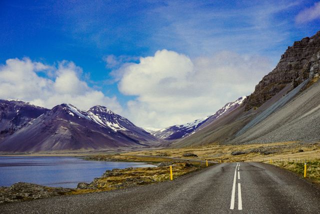 This scene captures a picturesque mountain road running alongside a tranquil lake under a vibrant blue sky with scattered clouds in Iceland. Ideal for use in articles or advertisements focusing on travel, road trips, adventure activities, or natural landscapes. Perfect for promoting Iceland as a travel destination or for inspiring wanderlust in readers.