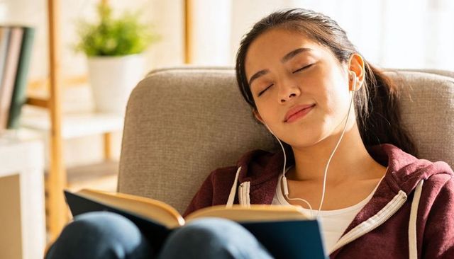 Young woman relaxing with book and earphones on sofa, cozy reading and listening at home