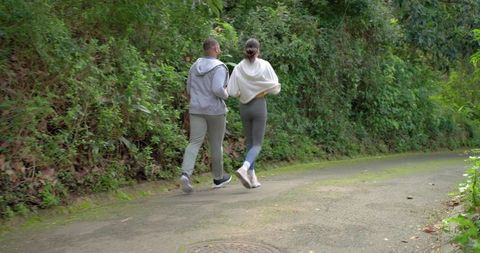 Diverse couple jogging on mossy paved park trail wearing hoodies and leggings for fitness