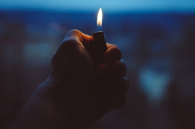Close-up of a hand holding a lighter with a flame, set against a dim background with twilight sky. The warm flame contrasts against the cooler blues of the twilight. Ideal for usage in themes related to fire safety, ignition, energy, nighttime activities, or concepts of illumination and warmth. Can also be used for illustrating themes of survival, camping, or exploring in darkness.