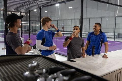Padel team taking coffee break at indoor purple court, teammates chatting and laughing