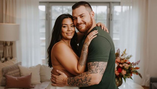 Smiling couple embracing in cozy sunlit living room with tattoos, ring and floral bouquet