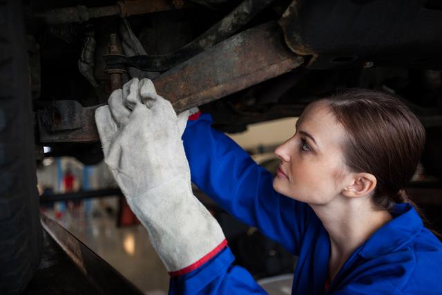 Female mechanic is focused on repairing a car in an auto garage. She is wearing a blue uniform and protective gloves, working diligently on the car's underside. Suitable for promoting gender diversity in engineering, automotive repair services, and technical skill workshops. Ideal for articles or advertisements about women in technical fields and professional training programs.