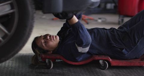 Auto mechanic lying on creeper inspecting vehicle undercarriage in garage workshop