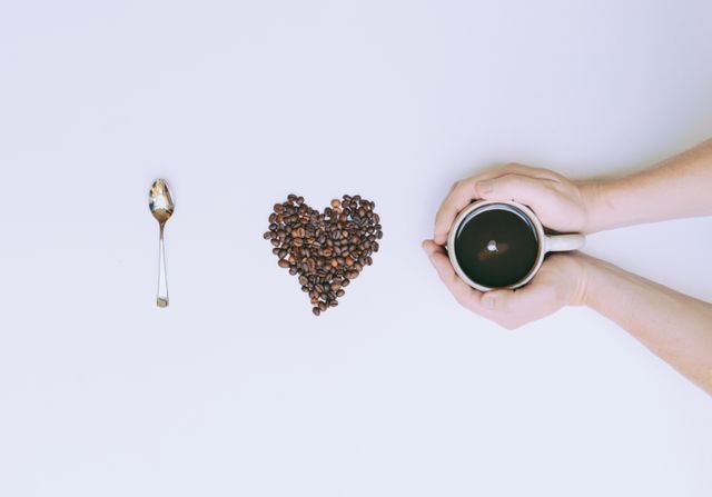 Hands holding coffee mug next to heart-shaped coffee beans and spoon with coffee residue. Ideal for promoting coffee brands, cafes, love for coffee messages, and minimalist aesthetic designs.