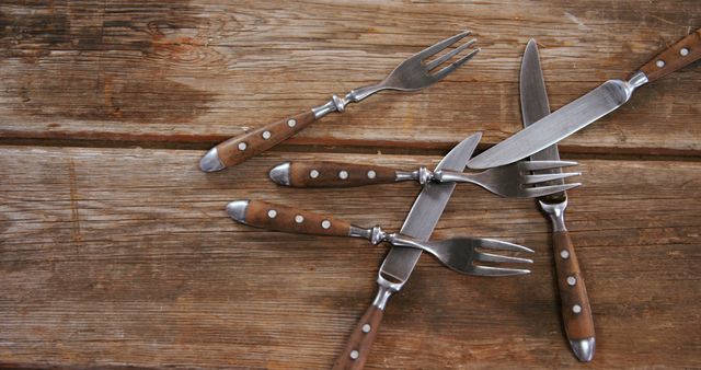 A set of forks and knives with wooden handles are arranged in a hashtag pattern on a rustic wooden table, with copy space. This arrangement showcases a creative presentation of cutlery, often used to enhance the visual appeal of dining settings.