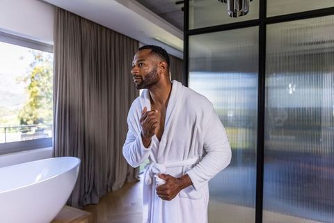 Mature african american man grooming face in white bathrobe beside freestanding bathtub
