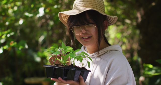 Young woman enjoying gardening holding a tray of seedlings in an outdoor garden. She wears glasses and a straw hat, dressed casually with a white hoodie. Ideal for content related to gardening, nature, environmental sustainability, and summer outdoor activities.