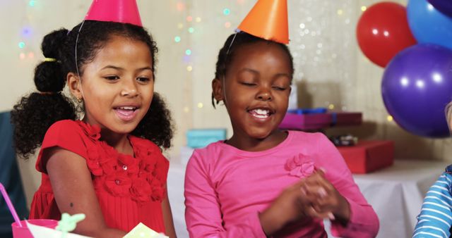 Two young African American girls celebrate at a birthday party, adorned with colorful hats and surrounded by balloons and gifts, with copy space. Their joyful expressions capture the excitement and happiness of a child's festive occasion.