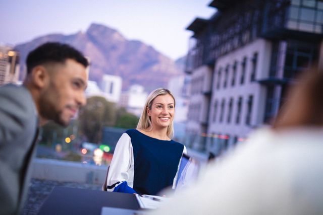 Businesswoman smiling during an outdoor meeting with a diverse colleague, set against a backdrop of city buildings and mountains. Ideal for use in corporate presentations, business websites, and promotional materials highlighting teamwork, diversity, and modern work environments.